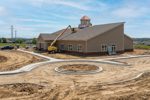 Beige one-story building under construction with a circular concrete driveway and a yellow boom lift on the roof edge, workers nearby in a sunny lot.