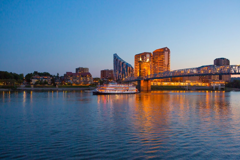 Covington-Kentucky - ABS Services Evening city skyline across a calm river, a lit steel bridge and illuminated buildings reflecting in the water.