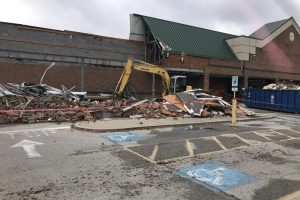 Yellow excavator amid a rubble pile outside a brick storefront, with a taped-off demolition zone.