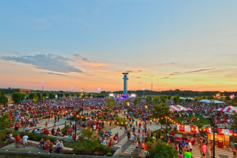 Large outdoor festival at sunset with a packed crowd, a central stage tower in the distance, and striped tents along a plaza area.