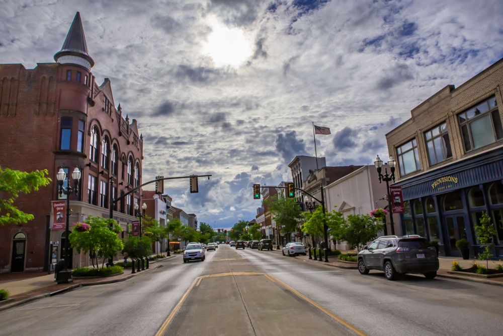 Downtown street with historic brick buildings, trees, and cars under a cloudy sky.