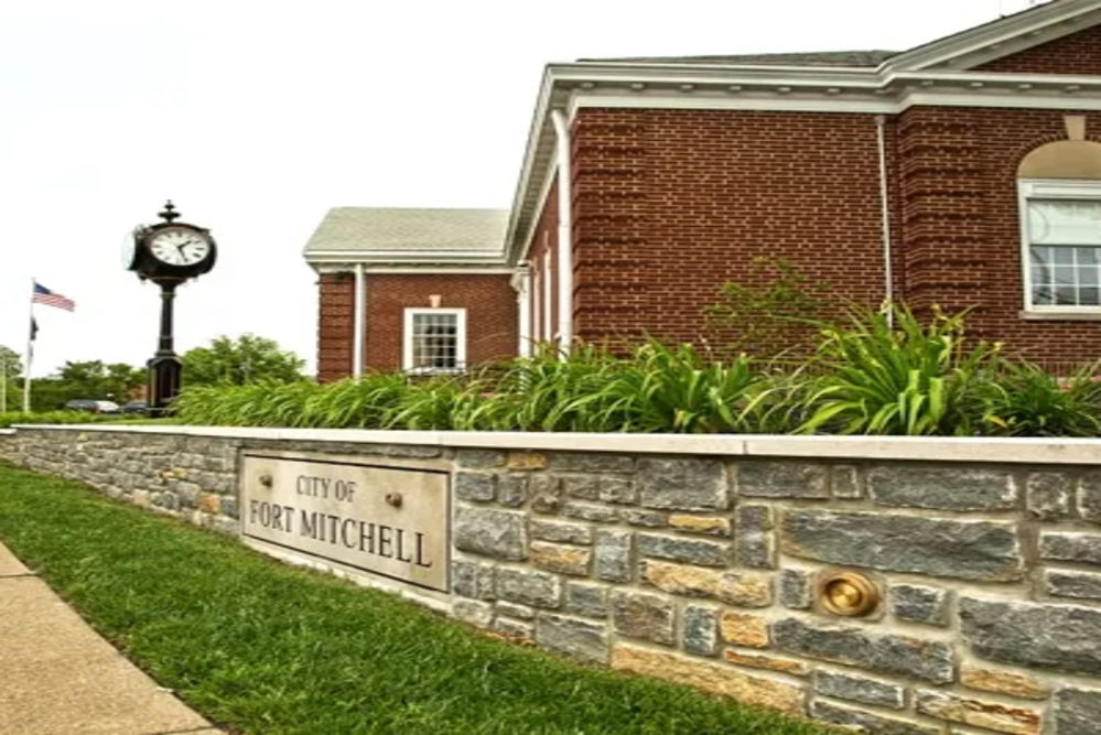 Fort Mitchell - ABS Services Stone wall with 'City of Fort Mitchell' plaque in front of a brick building and clock, with greenery and an American flag nearby.