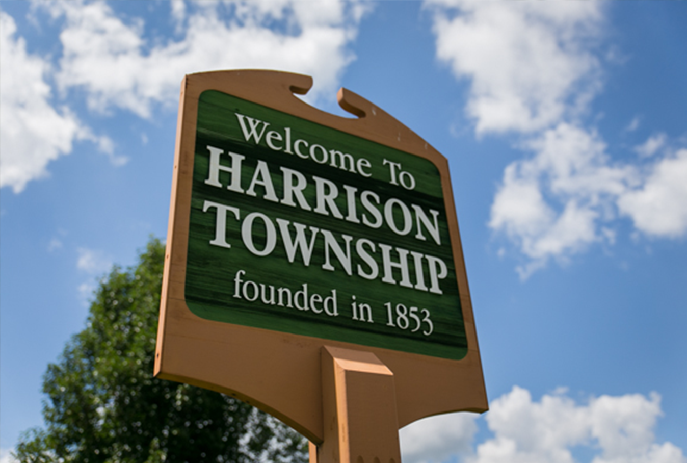 Green welcome sign reading 'Welcome To Harrison Township, founded in 1853' against a blue sky, outdoors image.