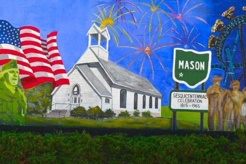Mural depicting a white church, waving American flag, green Mason sign, fireworks, and a statue trio on a roadside backdrop at dusk. (Celebration theme)