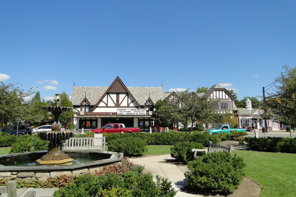 Mariemont - ABS Services Tudor-style storefronts line a sunny town square with a multi-tier fountain, benches, and neatly trimmed hedges in the foreground.